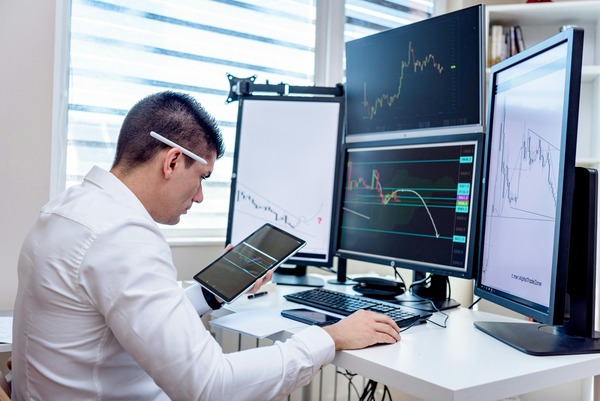 Trader reviewing several computer monitors with cryptocurrency price charts and a tablet