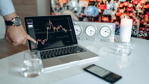Man pointing at a cryptocurrency price chart on a laptop screen next to a row of clocks